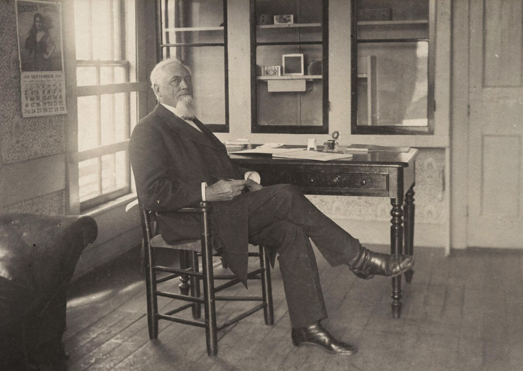Royal Rounsevel sits to the side of a desk, near a window. He is wearing a suit and has a white beard.