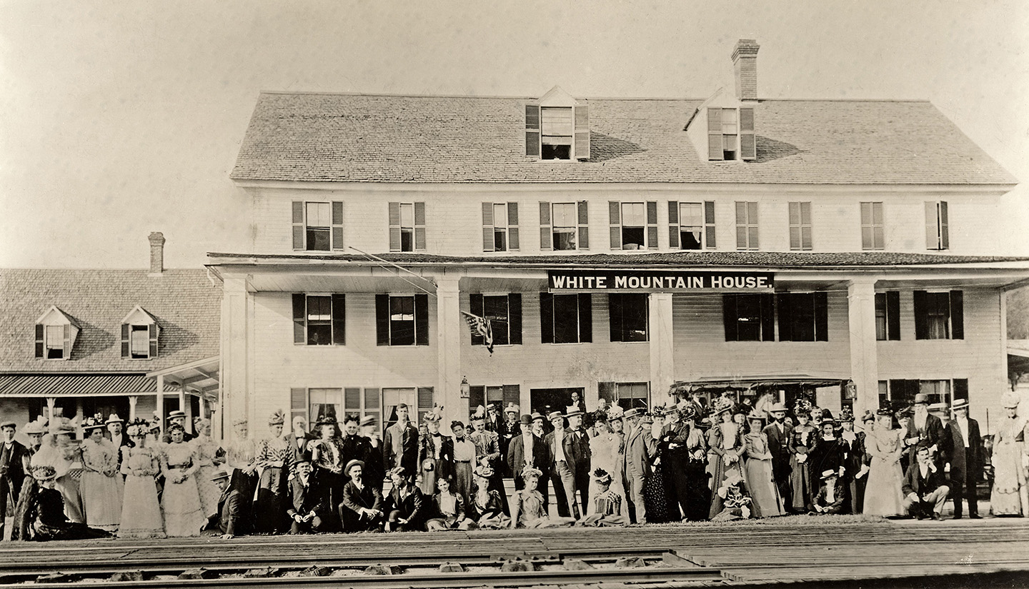 A railroad track is visible in front of a crowd of people in front of the White Mountain House, which has a wide front porch, a row of windows above the porch, and two dormers.
